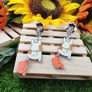 May include: Two pairs of cat-themed dangle earrings with a crab charm. The earrings are made of wood and metal. The cats are white with orange accents and are hanging upside down. The crabs are orange.