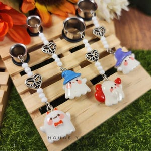 May include: A set of silver-toned ear gauges with dangling white beaded chains. Each chain features a filigree heart charm and a small, white ghost charm wearing a hat or bow. The text "HOUSE YEAGER" is visible.