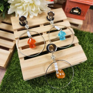 May include: Three pairs of silver-tone hoop earrings with mushroom charms in orange, blue, and yellow. Each earring has a silver-tone plug and is decorated with white beads and a mushroom pendant. The earrings are displayed on a wooden pallet.