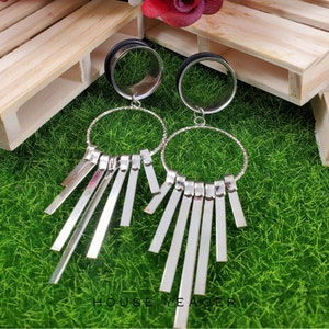 May include: A pair of silver hoop earrings with dangling silver bars. The earrings are hanging from a wooden surface with a green grassy background.