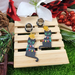 May include: A pair of black cat Christmas tree earrings with a star on top and colorful lights. The earrings are made of acrylic and are attached to silver metal plugs.