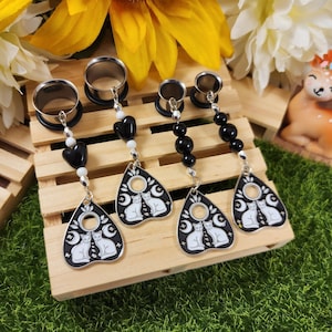 May include: Four pairs of silver-toned ear gauges with black and white beaded dangles. Each dangle features a black and white planchette design with two white cats, a crescent moon, and stars. The gauges are displayed on a wooden surface.