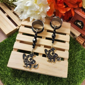 May include: A pair of black and gold witch-themed ear gauges. Each gauge features a black witch riding a broom, with a crescent moon and stars design. The gauges are connected to a black beaded chain and silver-colored tunnels.