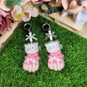 May include: A pair of pink cat earrings with white details and silver accents. The cats are wearing pink scarves and have pink hearts on their chests.