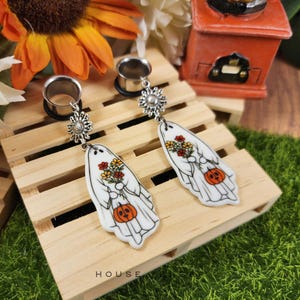 May include: A pair of silver-toned ear gauges with dangling white ghost-shaped charms. Each charm features a bouquet of flowers and a jack-o'-lantern. The word "HOUSE" is printed at the bottom. The gauges are on a wooden pallet.