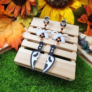 May include: A pair of Halloween themed earrings featuring a black and white ghost face knife charm, orange and black beads, and a white bow. The earrings are displayed on a wooden surface.