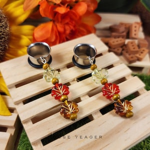 May include: A pair of stainless steel ear gauges with dangling leaf-shaped charms. The charms feature red, amber, and clear glass leaves, interspersed with wooden beads. The gauges are displayed on a wooden pallet.