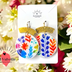 May include: A pair of colourful floral earrings with gold accents. The earrings feature a white background with blue, red, orange, and yellow floral designs. The earrings are hanging from gold hooks.