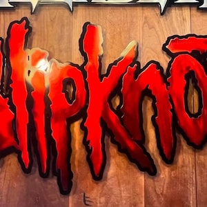 Puede incluir: Un letrero metálico rojo y negro con la palabra "Slipknot" en una fuente estilizada. El letrero tiene un efecto degradado, que pasa del rojo al naranja. El fondo es una superficie de madera.