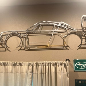May include: A silver metal wall sculpture of a sports car, mounted on a beige wall. The artwork shows the car's outline, including wheels, windows, and a rear spoiler. The metal has a polished finish, creating a modern aesthetic.
