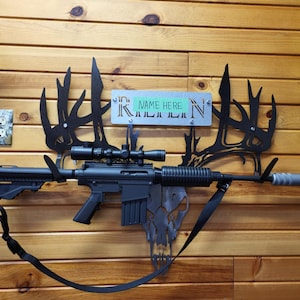 May include: A black rifle with a scope mounted on top is displayed on a wooden wall. The rifle is hanging from a metal deer antler silhouette. A metal sign with the text "NAME HERE" is attached to the antler silhouette.