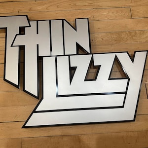 May include: A white and black sign with the words "THIN LIZZY" in a bold, stylized font. The letters are white with a black outline, set against a wooden background. The sign is likely a decorative item or a piece of band merchandise.
