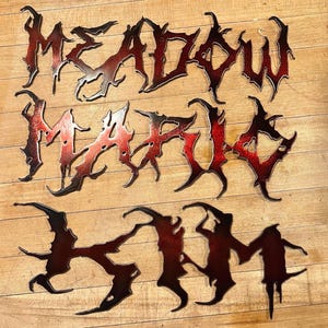May include: Three metal pieces spelling out "Meadow Marigolds" in a black and red gothic font. The metal is textured and has a red and black ombre effect.