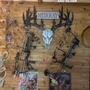 May include: A rustic wall display featuring a metal deer skull with antlers, a sign that reads "MURRAY", and two compound bows. The background is a wooden wall with additional hunting-themed decor. The text "HAMMERED BLACK AND SILVER" is at the top.