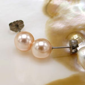 Könnte beinhalten: Ein Paar pfirsichfarbene Perlenohrringe mit silbernen Stiften und Verschlüssen. Die Ohrringe werden auf einem weißen und rosafarbenen Muschelhintergrund präsentiert.