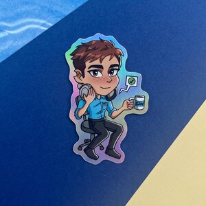 Może przedstawiać: Holograficzna naklejka przedstawiająca postać z kresk&oacute;wki z brązowymi włosami, ubraną w niebieską koszulę i czarne spodnie, trzymającą kubek i słuchawki. Na kubku widnieją litery "SDN". Nad głową postaci znajduje się dymek z ptaszkiem.