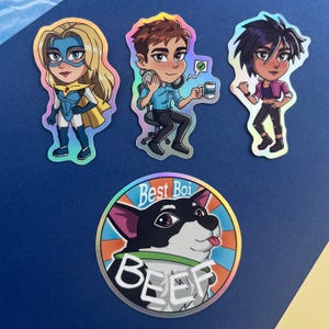 May include: A collection of holographic stickers featuring cartoon characters and a dog. The stickers include a superheroine, a person with headphones, and a person in a pink top. The dog sticker says "Best Boi" and "Beef".