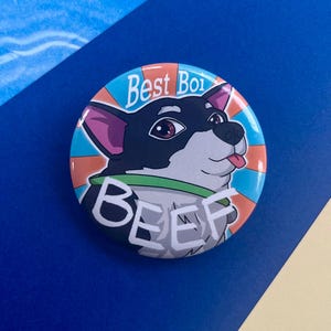May include: A round button with a cartoon dog illustration. The dog has black and white fur, pink ears, and a green collar. The button has the text "Best Boi" and "BEEF" in white.