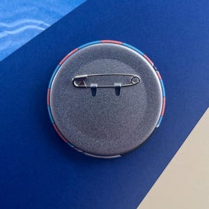 Może przedstawiać: Okrągła, srebrna przypinka z agrafką z tyłu. Krawędź przypinki ma wz&oacute;r w r&oacute;żowe, niebieskie i czarne paski. Przypinka jest umieszczona na niebieskim tle.