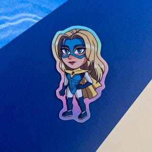Może przedstawiać: Holograficzna naklejka przedstawiająca kresk&oacute;wkową superbohaterkę z blond włosami, niebieską maską i niebiesko-białym kostiumem. Ma ż&oacute;łtą pelerynę i zdecydowany wyraz twarzy. Naklejka ma błyszczące wykończenie.