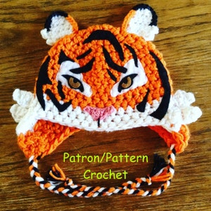 Puede incluir: Sombrero de tigre de crochet naranja, negro y blanco con un pompón blanco en la parte superior. El sombrero tiene una nariz de crochet negro y ojos marrones. El sombrero tiene una larga corbata de crochet negro, blanco y naranja.