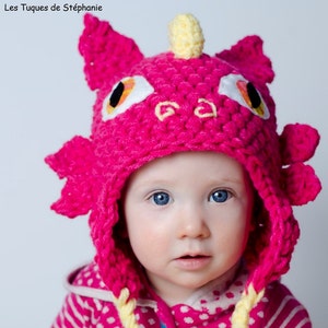 May include: A pink crocheted baby hat with a yellow unicorn horn and a yellow braid. The hat has a dragon design with orange eyes. The hat is on a baby wearing a pink and white striped shirt with white polka dots. The text "Patron/Pattern Crochet!!" is on the bottom of the image.