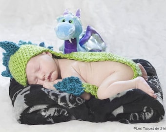 May include: A baby wearing a green and blue crocheted dragon costume sleeps on a black and white blanket with a blue plush dragon toy.