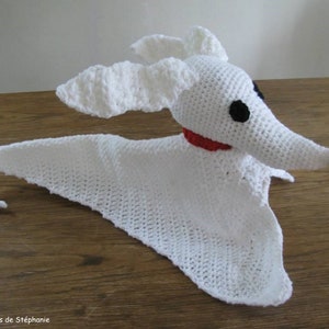May include: White crocheted ghost dog toy with black eyes and a red nose. The toy is lying on a wooden surface.