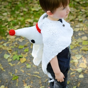 May include: A child wearing a white crocheted ghost costume with a red and black nose. The costume has a long tail and a hood. The child is standing on a path with fallen leaves.