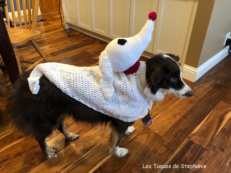 May include: A brown and white dog wearing a white crocheted ghost costume with a red nose and black eyes. The dog is standing on a wooden floor. The text "Les Tuques de Stephanie" is visible in the bottom right corner of the image.