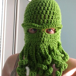 May include: A green crocheted hat with a face mask and long tentacles. The hat is made of a soft, textured yarn.
