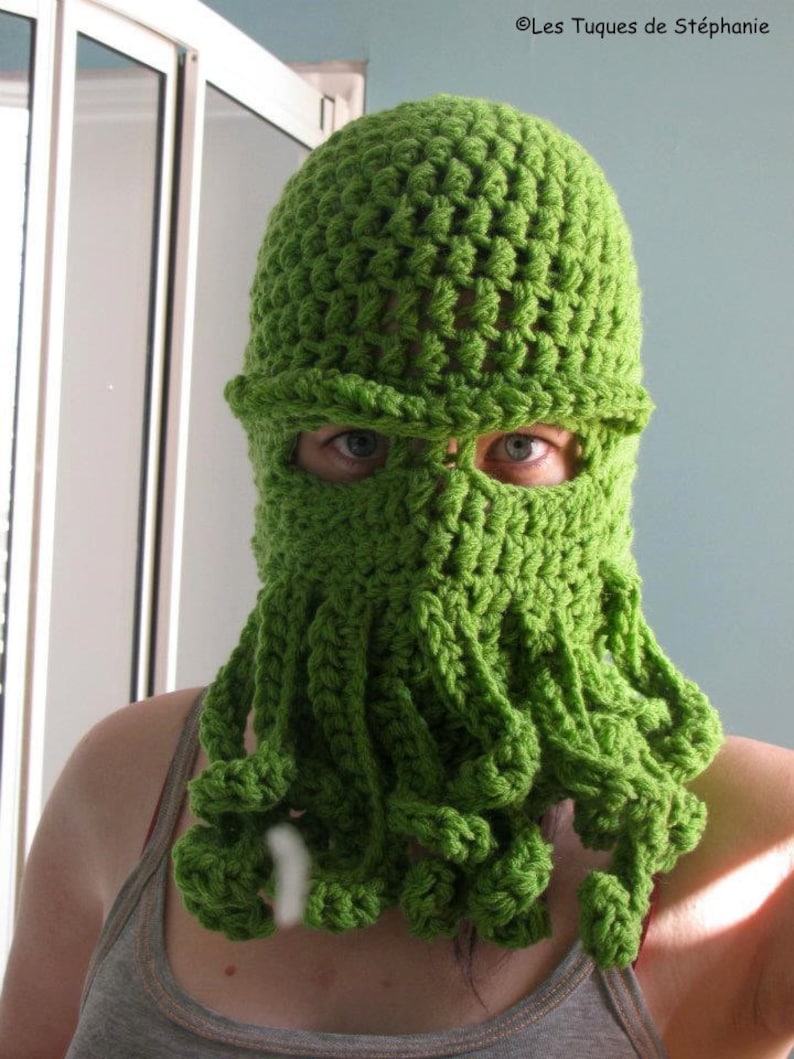 May include: A green crocheted hat with a face mask and long tentacles. The hat is made of a soft, textured yarn.