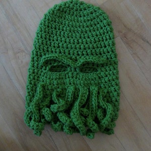 May include: Green crocheted hat with a face and tentacles. The hat has eye holes and a long, flowing beard of tentacles.