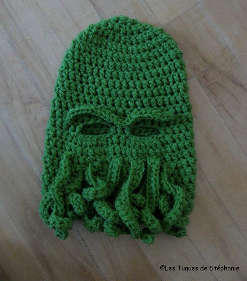 May include: Green crocheted hat with a face and tentacles. The hat has eye holes and a long, flowing beard of tentacles.