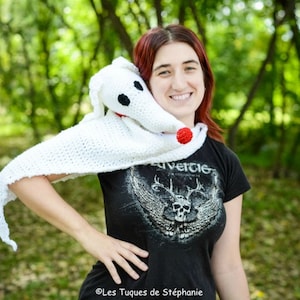 May include: A white crocheted shawl with a red nose and black eyes, resembling a ghost dog, is worn by a person with red hair. The person is wearing a black t-shirt with a skull and wings graphic and the text "Rivercie".