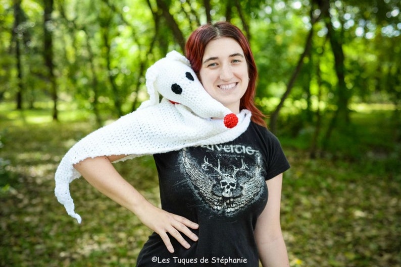 May include: A white crocheted shawl with a red nose and black eyes, resembling a ghost dog, is worn by a person with red hair. The person is wearing a black t-shirt with a skull and wings graphic and the text "Rivercie".