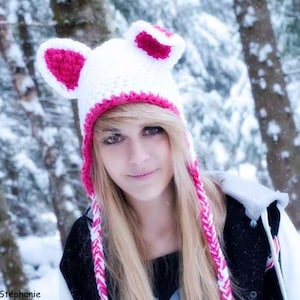 May include: A white crocheted hat with pink ears and a white and pink braid. The hat is worn by a person with long blonde hair. The background is a snowy forest.