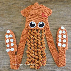 Puede incluir: Sombrero de calamar de crochet naranja con ojos blancos y círculos blancos en los tentáculos. El sombrero tiene una cola larga y está hecho de hilo.