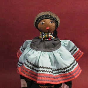 May include: A handmade doll with a dark complexion, wearing a light blue dress with orange and black trim. The doll has a beaded necklace and a woven head covering. The skirt is black with pink and blue geometric shapes.