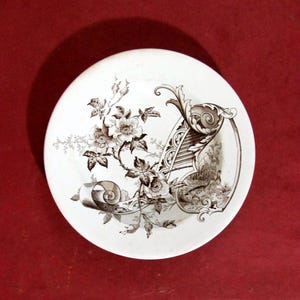 Puede incluir: Un plato de cerámica blanco con un diseño floral marrón. El plato presenta una ilustración detallada de flores, hojas y un pergamino decorativo. El diseño se encuentra sobre un fondo blanco, creando una estética vintage.
