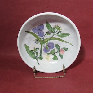 Puede incluir: Un cuenco de cerámica blanco con un diseño floral pintado a mano con flores moradas, verdes y rosas. El cuenco se exhibe sobre un soporte decorativo de metal sobre un fondo rojo. Es un objeto decorativo.