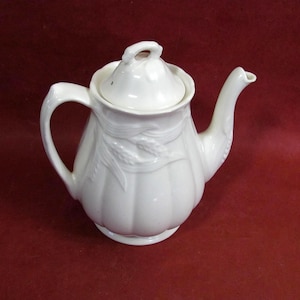 May include: A white ceramic teapot with a lid, handle, and spout. The teapot has a rounded body with vertical ridges and a wheat stalk design. The handle is curved, and the spout is slender. The teapot is on a red background.