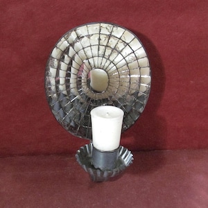 May include: A decorative wall sconce with a circular, mirrored design. The sconce features a small, white candle in a metal holder. The background is a deep red color.