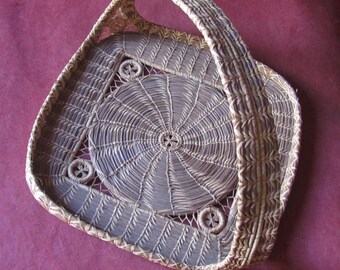 Puede incluir: Una cesta tejida con asa, hecha de materiales naturales. La cesta tiene un diseño circular en el centro y una forma cuadrada.