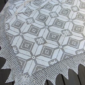 Pode incluir: Uma manta de crochê em tom branco sujo com um padrão geométrico repetitivo de diamantes e quadrados interligados. O tecido apresenta um design delicado de renda com uma borda pontiaguda e festonada que emoldura o trabalho de agulha.