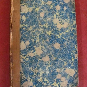 May include: A rectangular antique book with a marbled cover featuring a pattern of blue and yellow swirls. The spine and corners are covered in brown leather, showing signs of wear along the edges and the binding of the hard cover.
