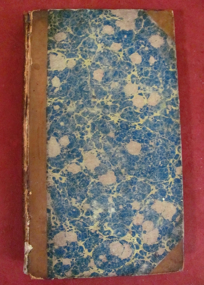 May include: A rectangular antique book with a marbled cover featuring a pattern of blue and yellow swirls. The spine and corners are covered in brown leather, showing signs of wear along the edges and the binding of the hard cover.