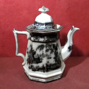 May include: An octagonal ceramic teapot featuring a white base with black transferware landscape patterns. The teapot includes a matching lid with a decorative finial, a curved handle, and a spout, all decorated with dark, detailed scenery.
