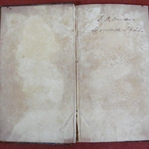 Op de afbeelding: Een open antiek boek met verouderde, gebroken witte pagina's die aanzienlijke vlekken en verkleuringen vertonen. De rechterpagina bevat handgeschreven cursieve inkttekst met de tekst E. A. Winder Woodside 1828, met een zichtbare rug.