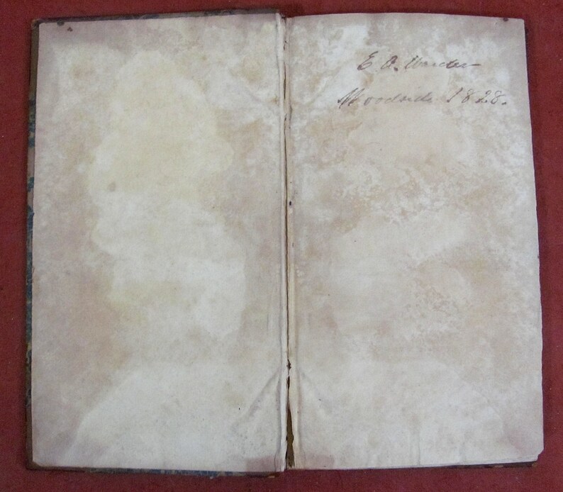 Op de afbeelding: Een open antiek boek met verouderde, gebroken witte pagina's die aanzienlijke vlekken en verkleuringen vertonen. De rechterpagina bevat handgeschreven cursieve inkttekst met de tekst E. A. Winder Woodside 1828, met een zichtbare rug.